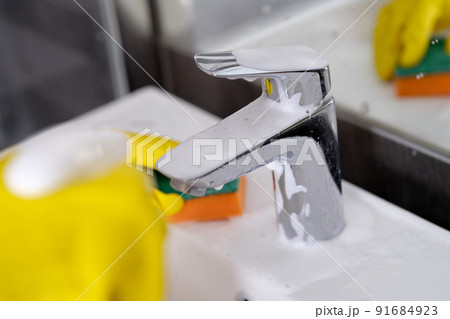 Hand in yellow rubber gloves holding sponge for washing dirty faucet with limescale 91684923