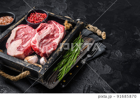 Fresh Raw Chuck roll, Tenders beef steaks in a wooden tray with herbs. Black background. Top view. Copy space 91685870