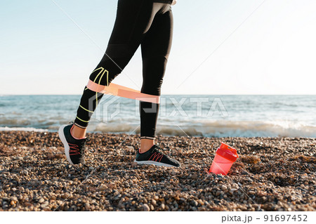 The concept of sport. A young woman exercises with a power band on her legs. There's a pink shaker on the ground. In the background, the sea and the horizon line. Close up 91697452