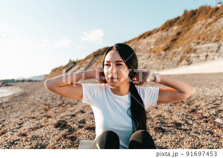 The concept of sport. Portrait of a young brunette woman shakes her abs sitting on a sports Mat. In the background mountains and wild beach 91697453