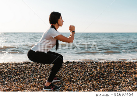 The concept of sport. Young brunette woman doing sports and squats. In the background, the sea and the shore. Copy space 91697454