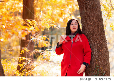 Elegant beautiful mature woman in a red coat posing by a tree. Autumn season. Copy space 91697479