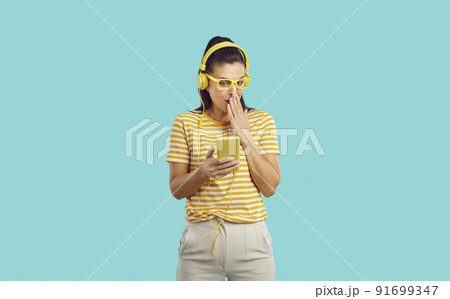 Young beautiful woman with headphones watching something incredible on the phone covers mouth in shock, looks shy, standing on blue background. Young beautiful woman with headphones watching something incredible on the phone covers mouth in shock, looks shy, standing on blue background. 91699347