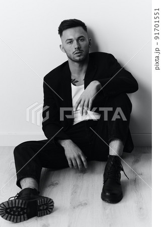 Modelling snapshots. Serious self-confident attractive handsome man in black jacket sitting on floor posing in white studio background. Black and White concept. Copy space Modelling snapshots. Serious self-confident attractive handsome man in black jacket sitting on floor posing in white studio background. Black and White concept. Copy space 91701551