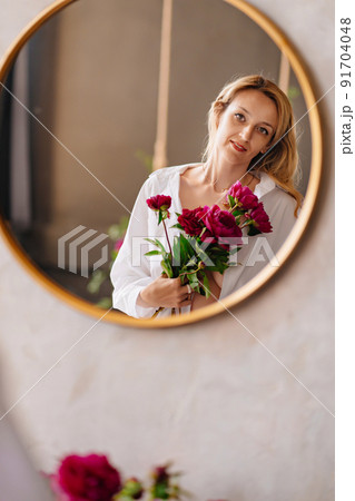 a blonde woman in a white shirt and a bouquet of peonies looks at in the mirror. a blonde woman in a white shirt and a bouquet of peonies looks at in the mirror. 91704048