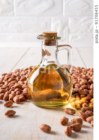 Peanut oil in a glass jug and raw peanuts on a white wooden table. 91706455