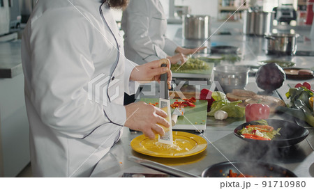 Male chef grating parmesan cheese on kitchen grater to cook gastronomy recipe in professional cuisine. Authentic cook preparing shredded cheddar ingredient to make gourmet meal dish. Male chef grating parmesan cheese on kitchen grater to cook gastronomy recipe in professional cuisine. Authentic cook preparing shredded cheddar ingredient to make gourmet meal dish. 91710980