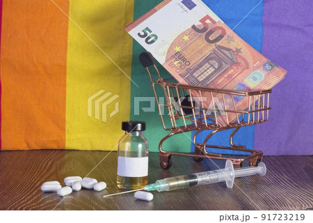 Injection, vaccine vial and medicinal tablets, Euro banknotes in a shopping cart. Background of a pride rainbow flag. 91723219