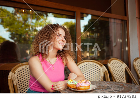 Portrait of beautiful curly young woman in cafe on summer terrace, delicious colorful donuts, sweet pastries. Lunch break, dreamy and happy. summertime 91724583