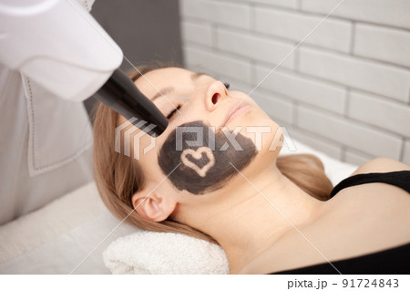 cosmetologist does procedure of carbon peeling of face. Black mask, laser skin cleansing. Modern treatments. Cuts heart. 91724843