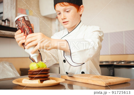 child boy in chef's costume prepares chocolate pancakes in kitchen in cafe. Pours chocolate. child boy in chef's costume prepares chocolate pancakes in kitchen in cafe. Pours chocolate. 91725613