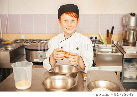 red-haired boy in chef's costume cooks pancakes in kitchen, kneads dough, assistant makes breakfast. Funny, breaks egg into bowl. smile 91725645