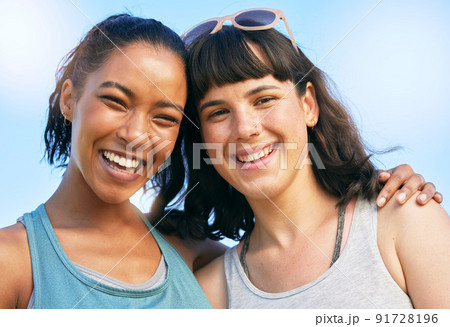 Portrait of two smiling friends embracing while standing together against blue sky. Smiling happy young women bonding and hugging outside over summer break. Close best friends having fun on a weekend 91728196