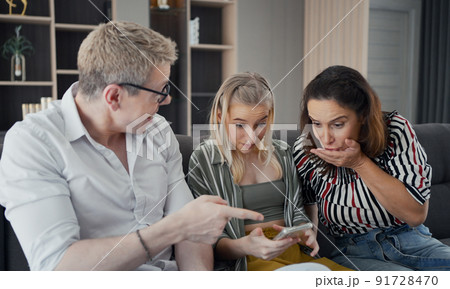 Caucasian parents sitting on sofa and excited daughter using smart phone, they're happy and wondering in something on screen with strange face in living room. Caucasian parents sitting on sofa and excited daughter using smart phone, they're happy and wondering in something on screen with strange face in living room. 91728470