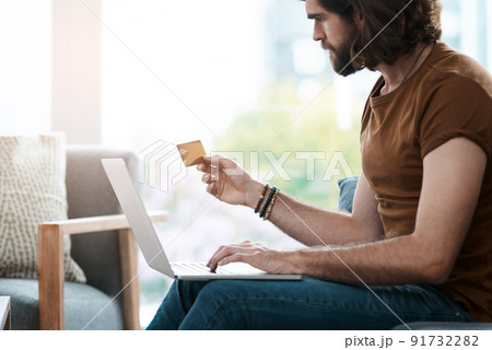 Time for some retail therapy. Shot of a handsome young man holding a credit card and using his laptop while relaxing in his living room. Time for some retail therapy. Shot of a handsome young man holding a credit card and using his laptop while relaxing in his living room. 91732282