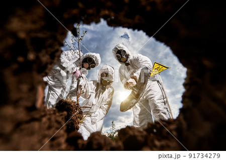 Concept of post apocalypse - people in white overalls and gas masks reforesting DEATH ZONE after hazard radioactive catastrophe on earth. Ecologists planting seedling. View from earth pit. 91734279