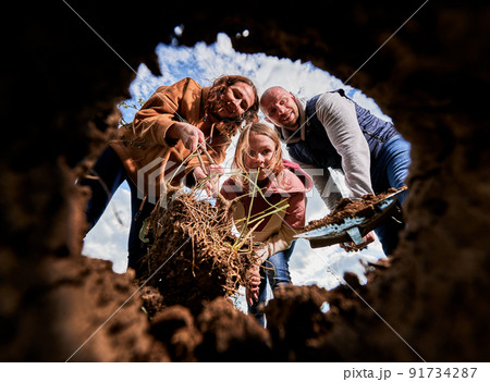Happy family planting tree sapling all together. Two adults, holding sapling and shovel, and school-age girl with joyful expression looking into dug hole for planting tree. View from inside pit. 91734287