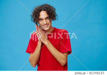 Sly curly tanned Caucasian guy in basic red t-shirt rubs his hands posing isolated on over blue background. Lifestyle and Emotions concept. Good offer with copy space for ad Sly curly tanned Caucasian guy in basic red t-shirt rubs his hands posing isolated on over blue background. Lifestyle and Emotions concept. Good offer with copy space for ad 91749137