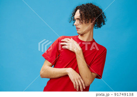 Unhappy discontented curly tanned Caucasian guy in basic red t-shirt looks with disgust aside posing isolated on over blue background. Lifestyle and Emotions concept. Good offer with copy space for ad Unhappy discontented curly tanned Caucasian guy in basic red t-shirt looks with disgust aside posing isolated on over blue background. Lifestyle and Emotions concept. Good offer with copy space for ad 91749138