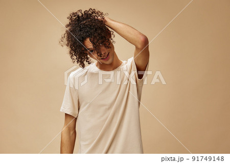 Funny awesome tanned curly man in basic t-shirt touches hair curls posing isolated on over beige pastel background. Fashion New Collection offer. People and Emotions concept. Free place for ad Funny awesome tanned curly man in basic t-shirt touches hair curls posing isolated on over beige pastel background. Fashion New Collection offer. People and Emotions concept. Free place for ad 91749148