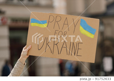 Cropped photo of a poster with phrase Pray for Ukraine on it in woman's hand. Somebody painted it in blue and yellow colors. Poster for supporting Ukraine. 91752633