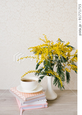 A beautiful composition of mimosa branches and a white tea cup with a saucer and a stack of notebooks on a table with a copy space A beautiful composition of mimosa branches and a white tea cup with a saucer and a stack of notebooks on a table with a copy space 91771755