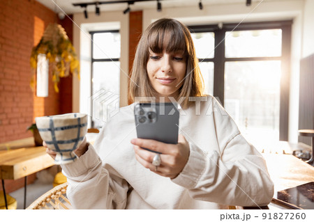 Woman with smart phone and coffee cup in sunny living room in the morning Woman with smart phone and coffee cup in sunny living room in the morning 91827260