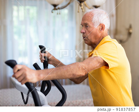 Elderly man exercising on elliptic trainer in living room Elderly man exercising on elliptic trainer in living room 91837857