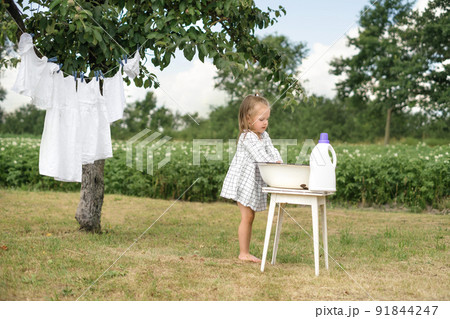 A barefoot girl in checkered dress washes children's underwear in basin in nature and hangs clean snow-white clothes on a clothesline to dry under a tree in the garden A barefoot girl in checkered dress washes children's underwear in basin in nature and hangs clean snow-white clothes on a clothesline to dry under a tree in the garden 91844247