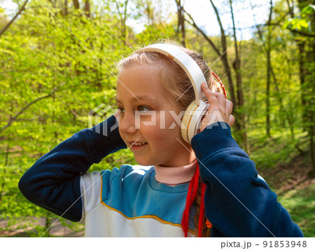 Adorable child girl in headphones listening to music using smartphone outdoor sunny day. Kid with gadget in the park. 91853948