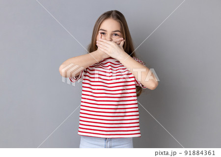 Portrait of dark haired little girl wearing striped T-shirt put hands on mouth, looking with fear in her eyes, keeping terrible secret. Indoor studio shot isolated on gray background. Portrait of dark haired little girl wearing striped T-shirt put hands on mouth, looking with fear in her eyes, keeping terrible secret. Indoor studio shot isolated on gray background. 91884361