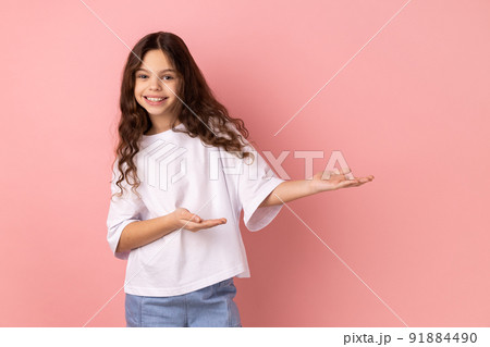 Portrait of kind-hearted generous little girl wearing white T-shirt showing welcome gesture and empty space on wall for your best advertising. Indoor studio shot isolated on pink background. Portrait of kind-hearted generous little girl wearing white T-shirt showing welcome gesture and empty space on wall for your best advertising. Indoor studio shot isolated on pink background. 91884490