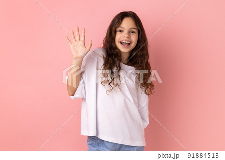 Portrait of friendly positive little girl wearing white T-shirt smiles toothily, raises palm greets friend, saying hello or goodbye. Indoor studio shot isolated on pink background. Portrait of friendly positive little girl wearing white T-shirt smiles toothily, raises palm greets friend, saying hello or goodbye. Indoor studio shot isolated on pink background. 91884513