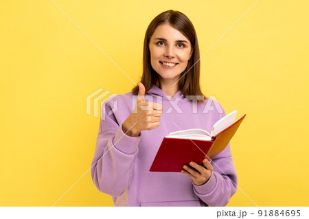 Portrait of happy satisfied woman showing thumbs up gesture holding and reading book, likes genre and interesting plot, wearing purple hoodie. Indoor studio shot isolated on yellow background. Portrait of happy satisfied woman showing thumbs up gesture holding and reading book, likes genre and interesting plot, wearing purple hoodie. Indoor studio shot isolated on yellow background. 91884695