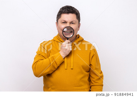 Portrait of middle aged man standing, holding magnifying glass on his teeth, looking with toothy smile, wearing urban style hoodie. Indoor studio shot isolated on white background. Portrait of middle aged man standing, holding magnifying glass on his teeth, looking with toothy smile, wearing urban style hoodie. Indoor studio shot isolated on white background. 91884941