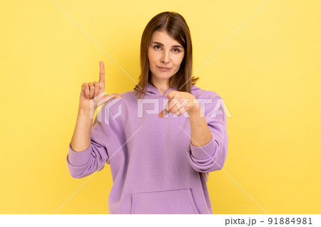 Portrait of frustrated young woman showing looser gesture and pointing at camera, looking with grumpy face, wearing purple hoodie. Indoor studio shot isolated on yellow background. Portrait of frustrated young woman showing looser gesture and pointing at camera, looking with grumpy face, wearing purple hoodie. Indoor studio shot isolated on yellow background. 91884981