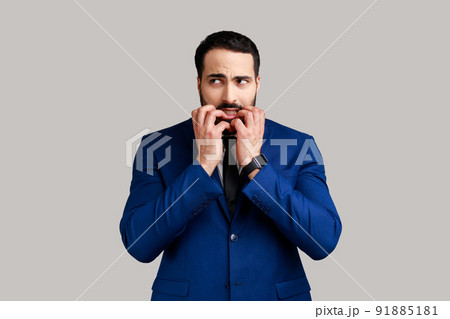 Bearded man biting nails, being nervous terrified, feeling frightened of challenge to start business, anxiety disorder, wearing official style suit. Indoor studio shot isolated on gray background. 91885181
