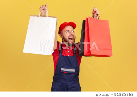 Portrait of satisfied bearded delivery man in blue uniform holding paper shopping bags, satisfied with black friday big sale, bringing order. Indoor studio shot isolated on yellow background. 91885268