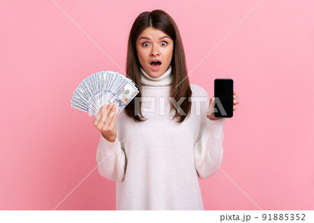 Shocked female holds big sum of dollars and showing smart phone with blank screen for advertisement, wearing white casual style sweater. Indoor studio shot isolated on pink background. Shocked female holds big sum of dollars and showing smart phone with blank screen for advertisement, wearing white casual style sweater. Indoor studio shot isolated on pink background. 91885352