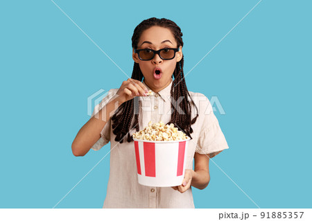 Excited woman with dreadlocks in 3d imax glasses watching movie film, hold popcorn, watching interesting movie, keeping mouth open, wearing white shirt. Indoor studio shot isolated on blue background. 91885357