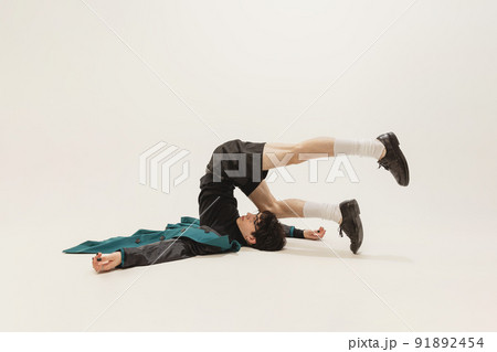 Portrait of stylish young man in black outfit and green coat posing, lying on floor isolated over grey studio background. Weirdness 91892454