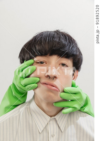 Portrait of young man with piercing posing with hands in rubber gloves on face isolated over grey studio background Portrait of young man with piercing posing with hands in rubber gloves on face isolated over grey studio background 91892520