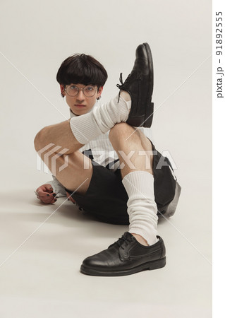Portrait of young man in black and white outfit sitting on floor, posing isolated over grey studio background. Piercing 91892555