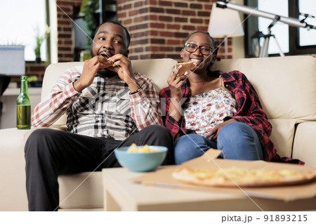 Young man and woman eating slice of pizza and drinking bottles of beer while they watch movie on tv together. Enjoying fast food takeaway delivery and beverage, watching television. 91893215