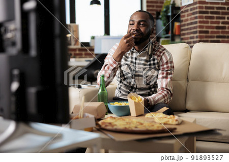 African american man eating chips while he watches film on tv, having fun with takeaway delivery and beer bottle. Watching movie on television and enjoying takeout fast food meal at home. 91893527