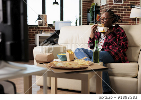 Happy woman using chopsticks to eat noodles from delivery box, enjoying meal in front of television watching movie. Eating fast food takeout package as dinner, looking at film on tv. 91893583