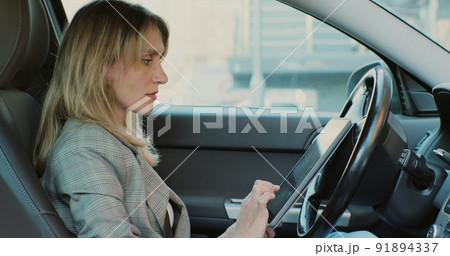 Young woman uses a program-navigator on the tablet, sits on the driver seat in the car. Woman working with tablet in car. Young woman uses a program-navigator on the tablet, sits on the driver seat in the car. Woman working with tablet in car. 91894337