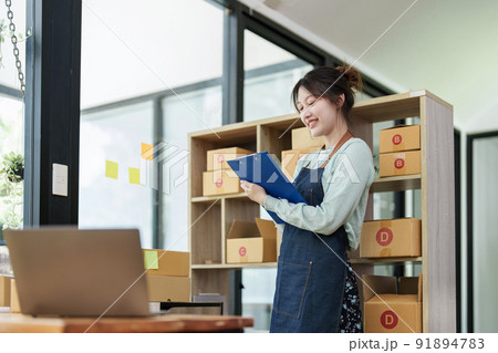 A portrait of a small startup, an SME owner, an Asian female entrepreneur checking orders to arrange the produce before packing the products in the inner boxes with the customers. Freelance concepts. A portrait of a small startup, an SME owner, an Asian female entrepreneur checking orders to arrange the produce before packing the products in the inner boxes with the customers. Freelance concepts. 91894783