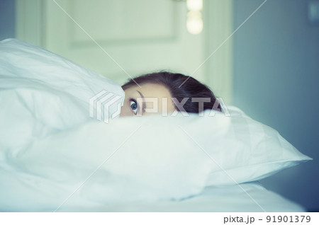 A young woman with huge frightened open eyes lying in bed and covers herself with the sheets and hides. Nightmares, insomnia, phobias, panic attacks. Concept 91901379