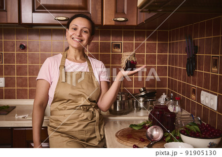 Charming pleasant woman confectioner holds a homemade cherry confiture jar, standing at cozy rustic kitchen interior Charming pleasant woman confectioner holds a homemade cherry confiture jar, standing at cozy rustic kitchen interior 91905130
